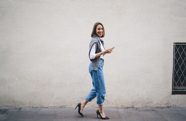 Side full length portrait of cheerful Caucasian woman dressed in stylish streetwear holding modern cellphone gadget and smiling at camera while walking near urban wall, happy female blogger laughing