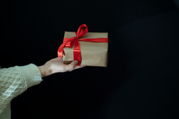 Female hands in a beige knitted sweater hold a gift wrapped in craft paper with a wide red ribbon on a black background. Holidays.