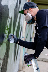 Graffiti artist in action, drawing on the wall with aerosol spray paint in a can, wearing protective face mask / respirator with filters. Street art culture concept. Vertical orientation.