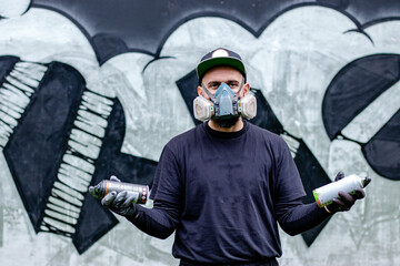 Obraz premium Graffiti artist posing in front of his drawing on the wall, with two aerosol spray paints in a can, wearing protective face mask / respirator with filters. Street art culture concept.