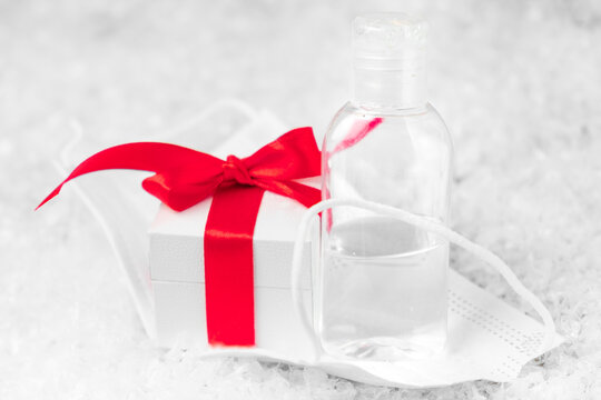 Gift box with a red bow from ribbon, sanitizer bottle in the protective face mask against coronavirus on the background of presents under Christmas tree. Christmas and New Year holidays during - Powered by Adobe
