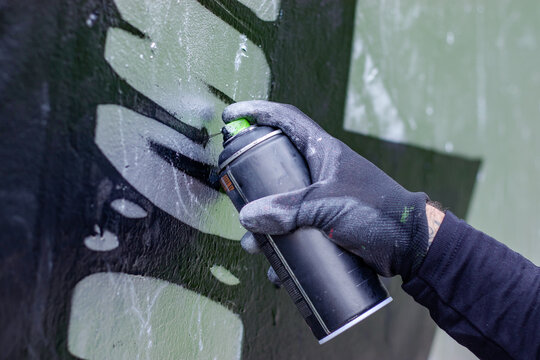 Hand of a graffiti artist in action, drawing on the wall with aerosol spray paint in a can, wearing protective gloves. Street art culture concept. Close up.