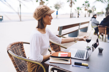 Positive caucasian female holding glass of wine resting after working day on cafe terrace using...