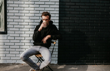 Fototapeta na wymiar A young charismatic guy, a hipster, drinks coffee against the background of a brick wall. Dressed in a black hoodie.