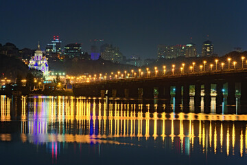 Picturesque panoramic photo of illuminated Paton Bridge over Dnipro River. Skyscrapers on the hills in the background.  City lights are reflecting on smooth water. Kyiv at autumn night, Ukraine