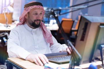 Handicapped Arabic businessman in tradition suit working in office during coronavirus  pandemic. Disabled businessman in the wheelchair works online in the office at the computer.