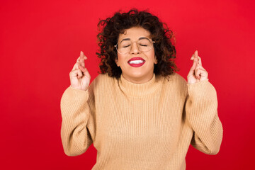 Joyful Young beautiful Arab woman wearing beige sweater against red background clenches teeth, raises fingers crossed, makes desirable wish, waits for good news, I have to win.