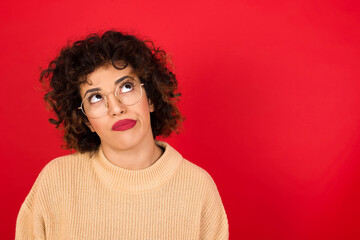 Young beautiful Arab woman wearing beige sweater against red background has worried face looking up lips together, being upset thinking about something important, keeps hands down.