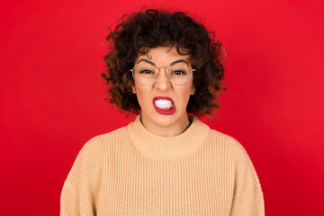 Mad crazy Young beautiful Arab woman wearing beige sweater against red background clenches teeth angrily, being annoyed with coming noise. Negative feeling concept.