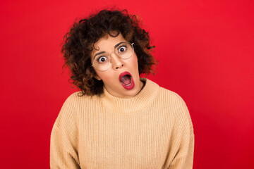 Expressive facial expressions. Shocked stupefied Young beautiful Arab woman wearing beige sweater against red background, keeps jaw dropped feels stunned from what he sees aside.