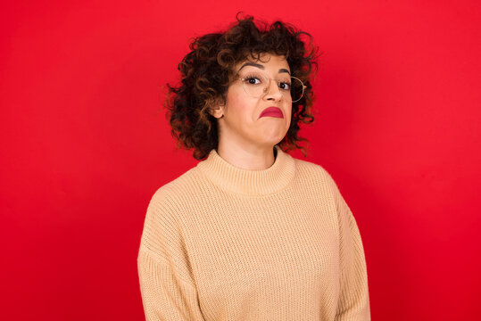 Young beautiful Arab woman wearing beige sweater against red background with snobbish expression curving lips and raising eyebrows, looking with doubtful and skeptical expression, suspect and doubt.