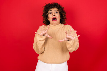 Dissatisfied Young beautiful Arab woman wearing beige sweater against red background frowns face, has disgusting expression, shows tongue, expresses non compliance, irritated with somebody.