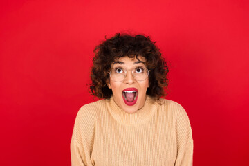 Surprised Young beautiful Arab woman wearing beige sweater against red background, shrugs...