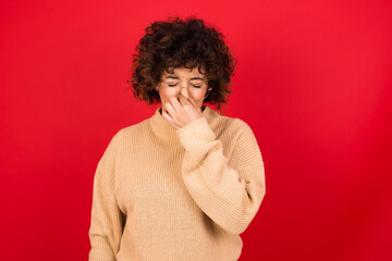 Fototapeta na wymiar Young beautiful Arab woman wearing beige sweater against red background, holding his nose because of a bad smell.