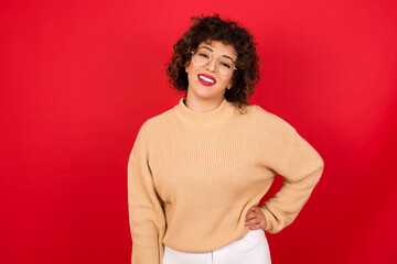 Studio shot of cheerful Young beautiful Arab woman wearing beige sweater against red background keeps hand on hip, smiles broadly.