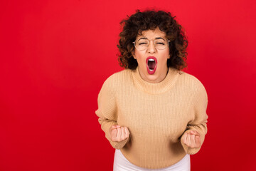 Joyful excited lucky Young beautiful Arab woman wearing beige sweater against red background cheering, celebrating success, screaming yes with clenched fists