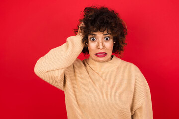 Portrait of confused Young beautiful Arab woman wearing beige sweater against red background holding hand on hair and frowning, panicking, losing memory. Worried and anxious can not remember anything.