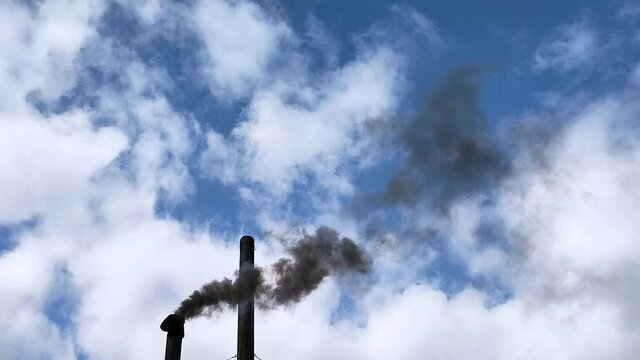 View of two chimneys against a blue sky with white clouds from which black smoke comes out.