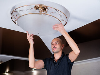 A young man is installing a plafond on a ceiling chandelier in the hall, standing on a stepladder