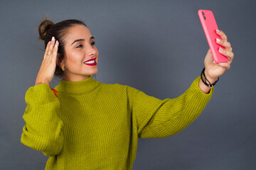 Young beautiful hispanic woman wearing green sweater against gray background smiling and taking a...