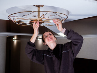 A woman stands on a metal staircase and changes the light bulb in the chandelier.