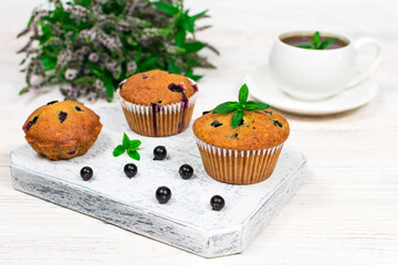 Cupcakes with black currants on a white wooden cutting board against the background of a cup and mint flowers.