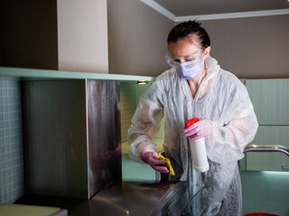 Woman in protective mask and glasses washes kitchen hood and furniture in the kitchen with disinfectant