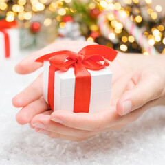 Close up woman hand holding a white gift box with a red ribbon on the background of presents, candy stick, Christmas tree and golden lights