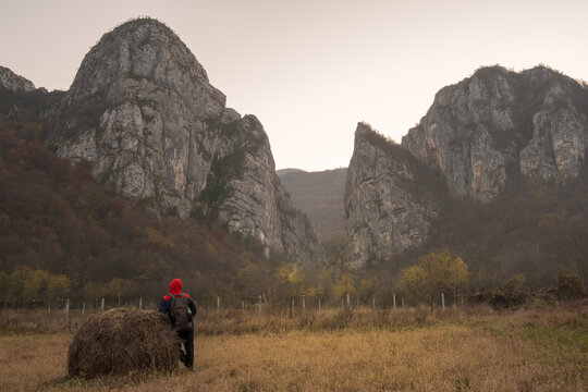 Mountain hiker in red jacket with hood looking at impressive mountain cliffs at the misty entrance of Jerma river canyon and foreground hay bale on a field - Powered by Adobe