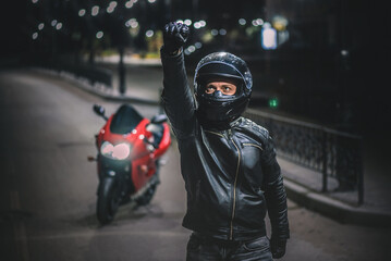 Motor biker is standing among the night road with raised up fist.