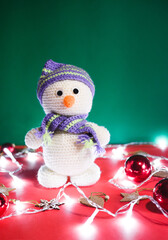 Cute handmade knitted white snowman with a carrot instead of a nose on a red and green Christmas background with decorations and a garland. A charming gift in the form of a small snowman toy.