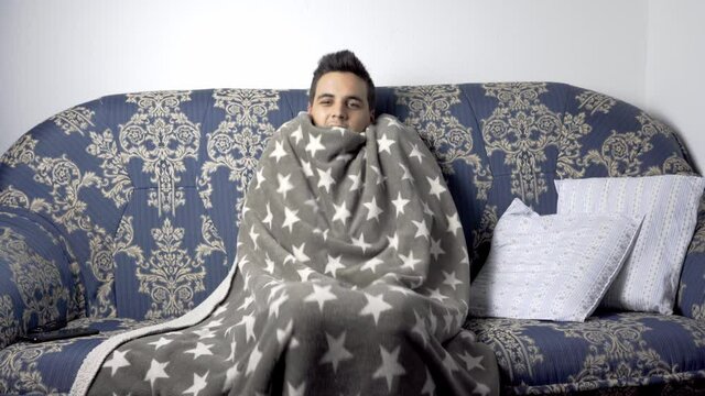 dark-haired teenager with black hair and a black sweatshirt sitting on a blue sofa with gold print and covering himself with a gray blanket. young latin man in winter in his living room