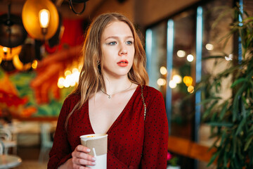 Young blonde woman holding a paper cup of hot coffee of takeaway in her hands indoors in cafe and looking in camera. copyspace. Coffee shop advertisement. Place for your text, concept business drinks