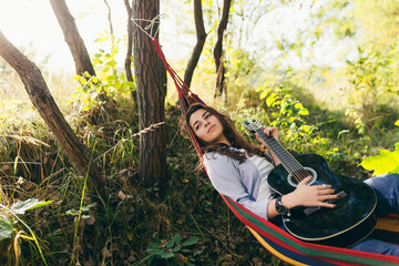 beautiful girl resting in a hammock in the middle of trees in the park, holding a guitar