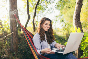 Young beautiful student is resting sitting on a hammock in the park and working on a laptop