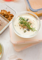Batch cooking scene with glass and plastic jars with prepared food inside. A glass jar of aromatic asparagus cream soup at the top of a pile.