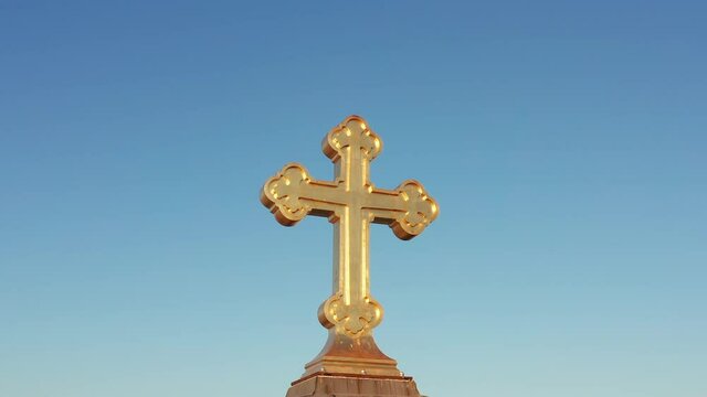 Shiny golden cross against clear blue sky. Orthodox christian crucifix illuminated by sunlight. Religious symbol of the faith in god made of gold. 