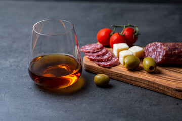 glass of whiskey and snacks sausage cheese olives and tomatoes on dark gray cement background