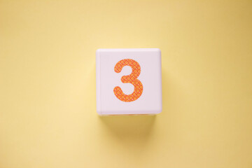 Close-up photo of a white plastic cube with a orange number 3 on a yellow background. Object in the center of the photo