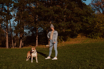 Full length of a beautiful smiling teenage girl with a hat and denim jacket posing while holding the leash of her dog breed American Staffordshire Terrier. Autumn walk