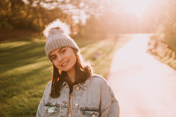 Portrait of a smiling beautiful teenage girl with a hat in a denim coat standing in the park. Sunny autumn day in nature