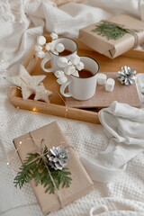 Naklejka premium Hot cocoa with marshmallow in a white ceramic mug, on a wooden tray and wrapped boxes. The concept of cosy holidays and New Year. Selective focus