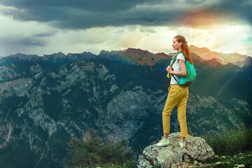  portrait of a modern woman tourist with a backpack standing on a rosk and looking around of landscaping views on background of the mountain range, cloudy sky and sun lights.