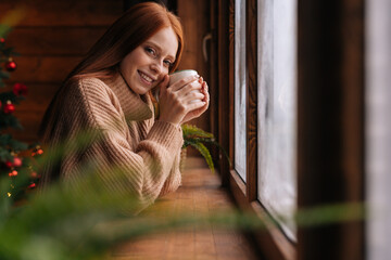 Portrait of cheerful young red-haired woman with cup standing by window at home at Christmas time....