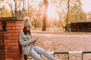 A young modern caucasian girl with a hat dressed in jeans sits in the park and uses the phone. Hello November