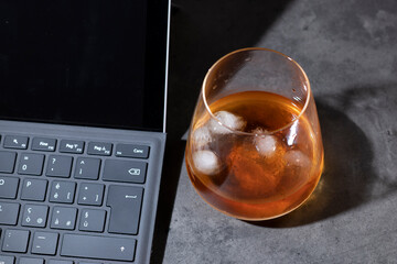 whiskey glass with ice next to laptop on dark gray cement background