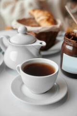 On the table white cup, chocolate cream, teapot. In the background, a basket of croissants