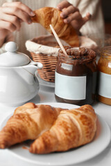 Two croissants on a white plate on the table. In the background, two jars of jam and a girl with a croissant in her hands