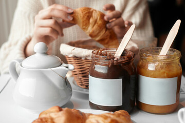 Two croissants on a white plate on the table. In the background, two jars of jam and a girl with a croissant in her hands