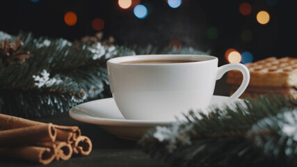 White cup of hot coffee on a brown wooden table. Green spruce branches, homemade waffles with souffle and cinnamon. Christmas lights and multicolored garland on background. New Year and Christmas